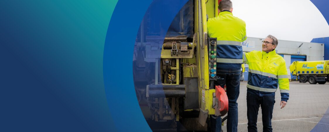 Twee mannen in gele en blauwe werkjassen van Rd4 staan bij een vuilniswagen op een bedrijventerrein. Eén man staat op de treeplank van de wagen, terwijl de ander met hem praat en naar hem lacht. Op de achtergrond zijn meer vuilniswagens en een gebouw met het Rd4-logo zichtbaar.