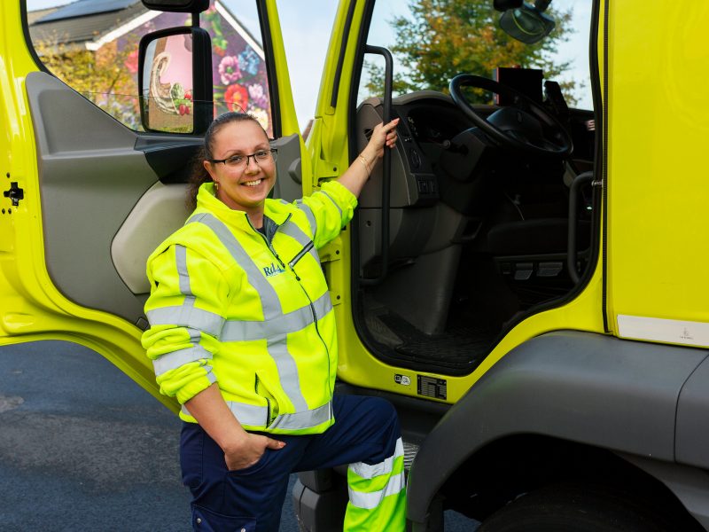 Een lachende inzamelmedewerkster staat naast de inzamelvrachtwagen.